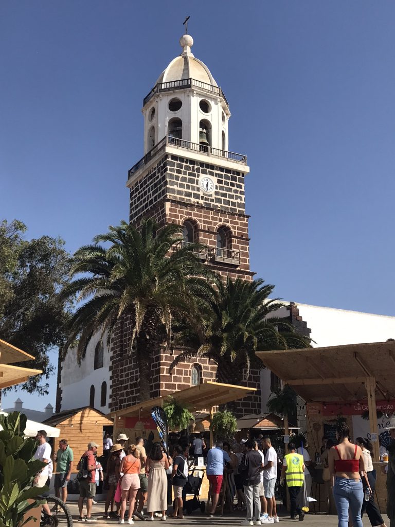 Clock tower at Teguise town, with sunny blue skies behind, gorgeous palm trees and a market stall at the base with people browsing