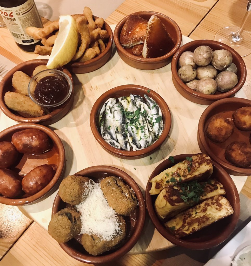 Picture of a selection of Tapas in small terracotta ramekins, there is some deep fried fish, anchovies, croquettes, potatoes, tortilla and chorizo